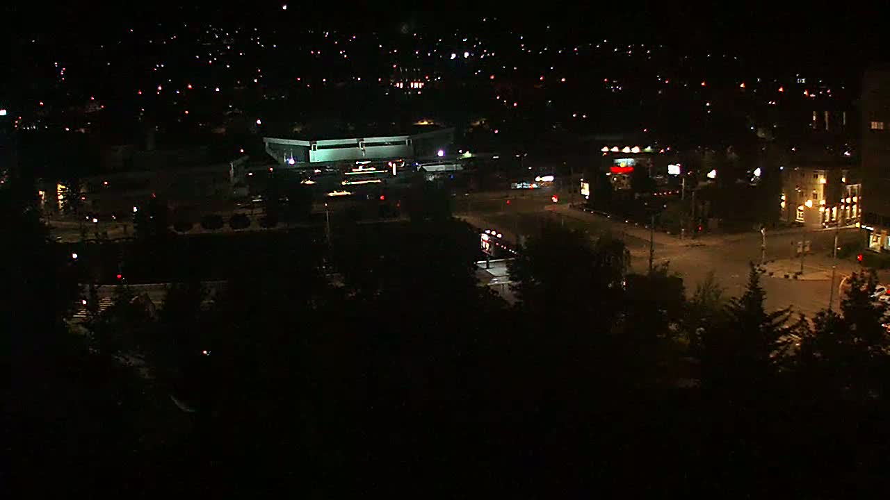A nighttime aerial view of a city with a large building, roads, and sparsely lit residential areas.