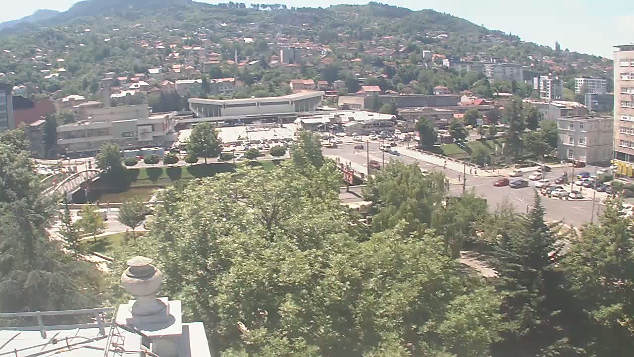 A high-angle view of a town nestled against a hillside on a sunny day shows buildings, roads, a river, and green spaces.