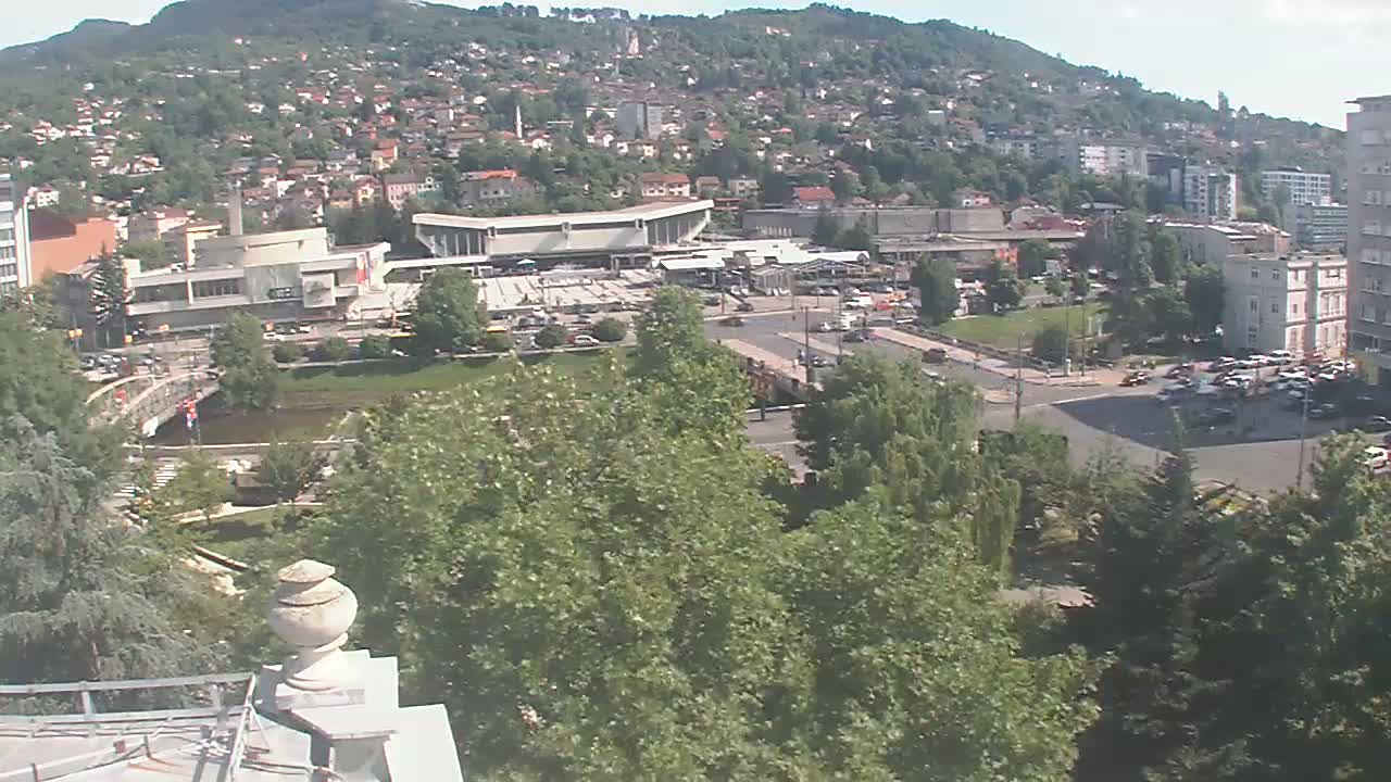 A high-angle view on a sunny day shows a city nestled against a hill, with a river flowing through a park in the foreground, and many buildings and trees visible.