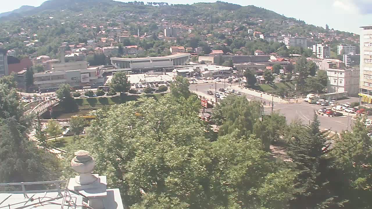 A high-angle view on a sunny day shows a town nestled in a hillside, featuring a large plaza with buildings, parked cars, and lush green trees surrounding a river.