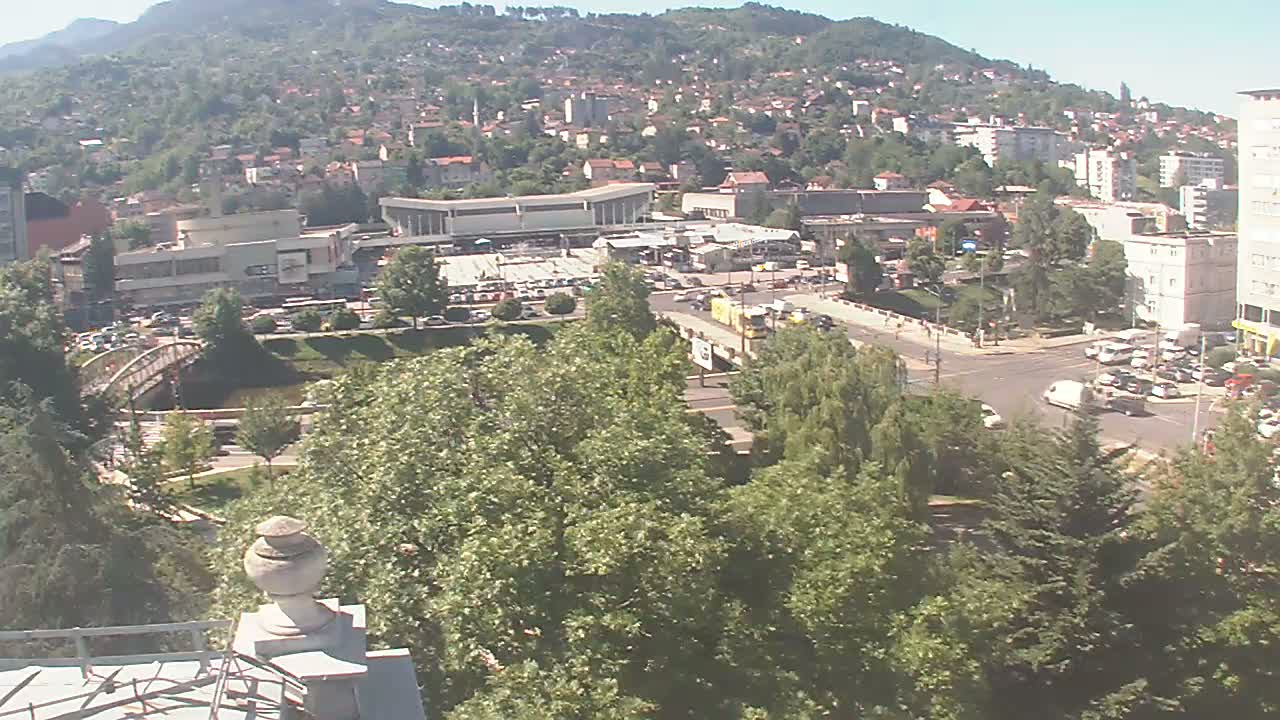 A high-angle, long shot on a sunny day shows a city nestled against a hill, featuring a large building complex, a bridge, a road intersection, and lush green trees.
