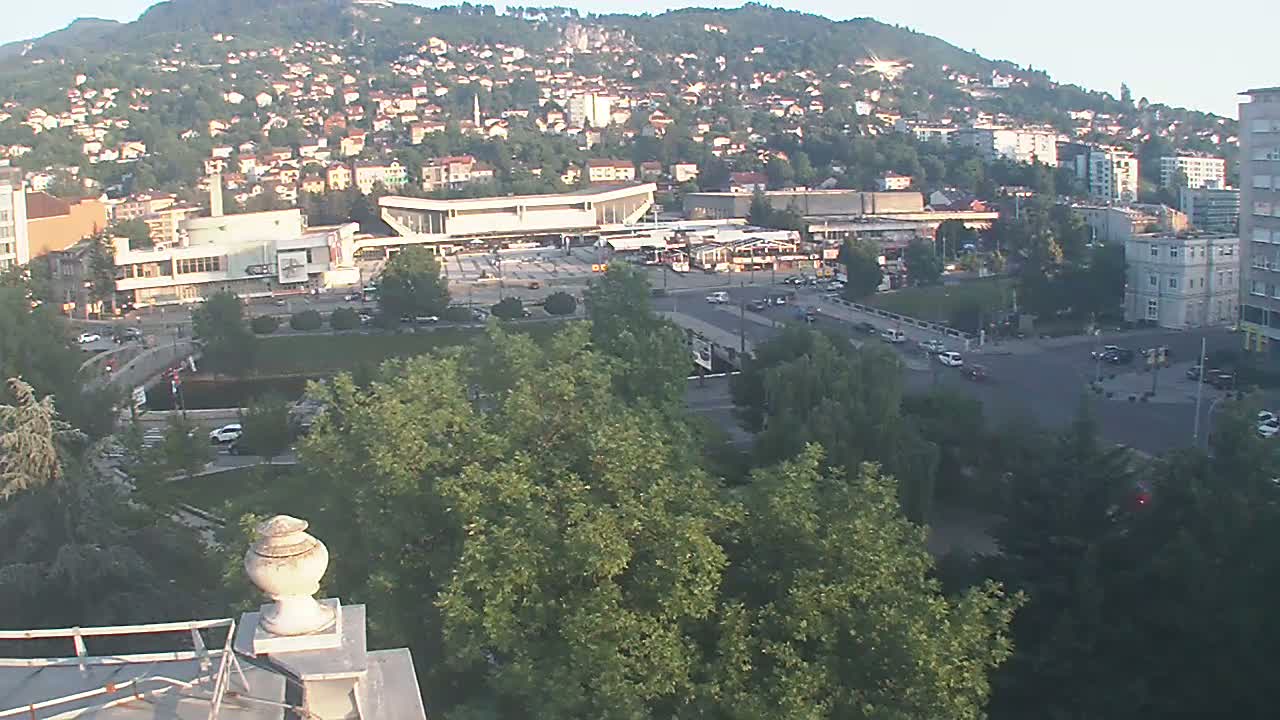 A sprawling city nestled against a hill, under a clear, bright sky, is viewed from an elevated perspective, showing a large plaza with a building complex and surrounding greenery.