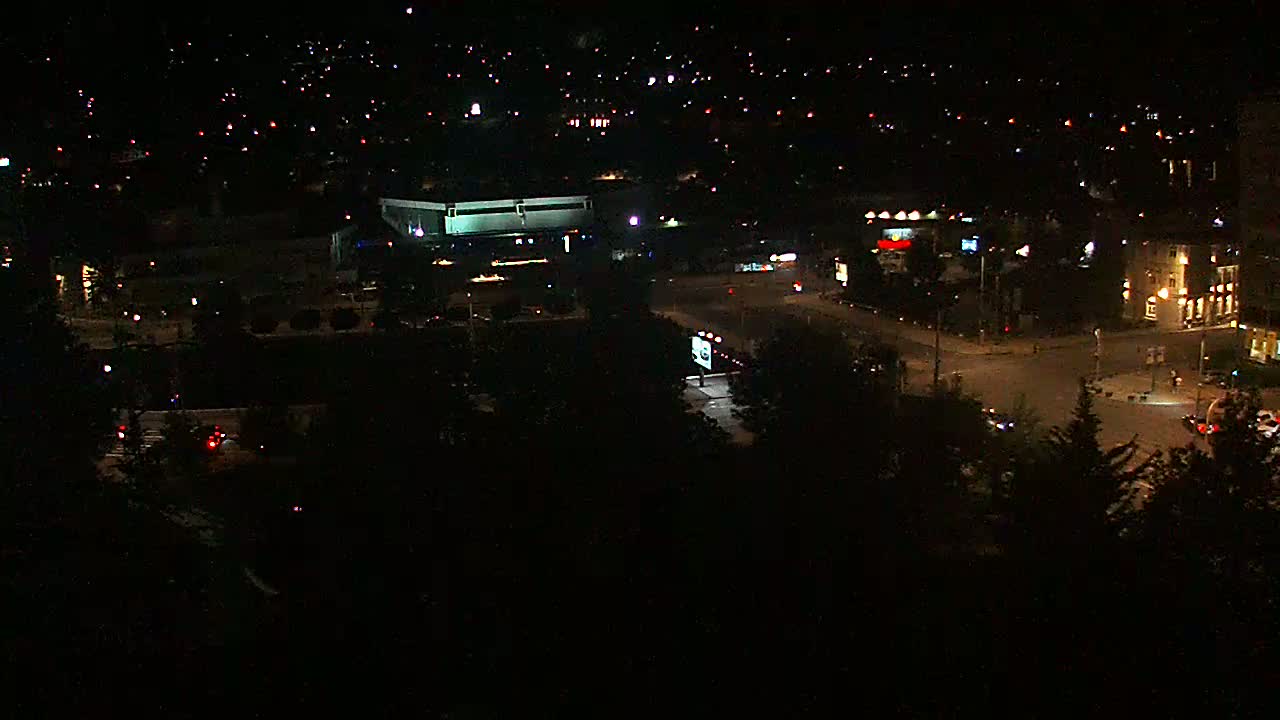A nighttime aerial view shows a city illuminated by streetlights and building lights, with cars driving on roads that intersect near a large building.
