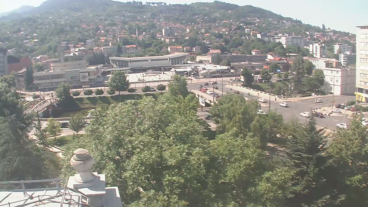 A high-angle view of a city nestled against a hillside on a sunny day, showing buildings, roads, trees, and a bridge over a waterway.