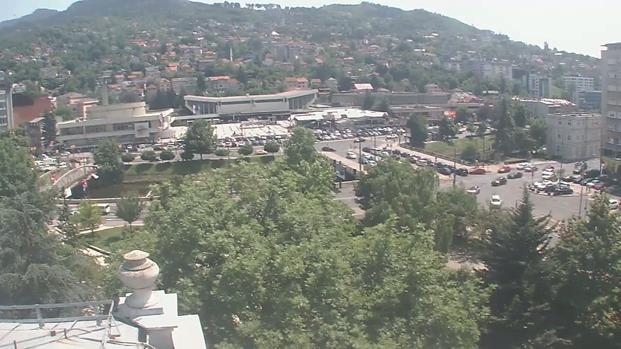 A sprawling town nestled against a hillside under a clear, sunny sky, features a large building complex, a small body of water, and numerous vehicles parked and driving on the roads.