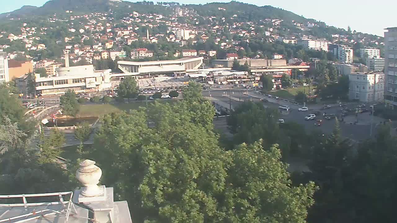 A high-angle, long shot on a sunny day shows a city sprawled across a hillside, with a large building and a plaza in the foreground, surrounded by lush green trees and parked cars.
