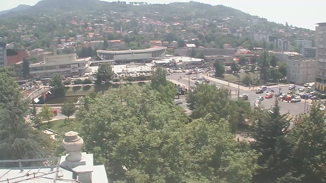 A high-angle view on a sunny day shows a city nestled in a valley beneath a hill, with a large building complex and numerous cars visible in the foreground.