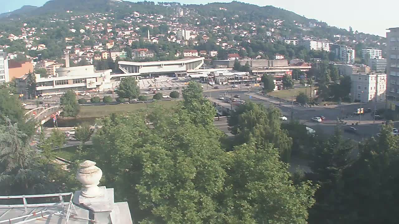 A high-angle, long shot on a clear day shows a city nestled in a valley at the foot of a hill, with a large building in the mid-ground, and lush green trees in the foreground.