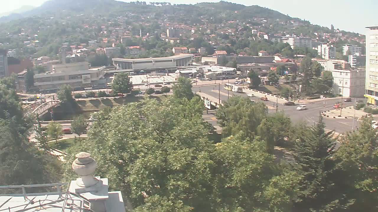 A high-angle view of a city nestled in a hillside on a sunny day, showing a large plaza, buildings, and green spaces with a bridge over a waterway.