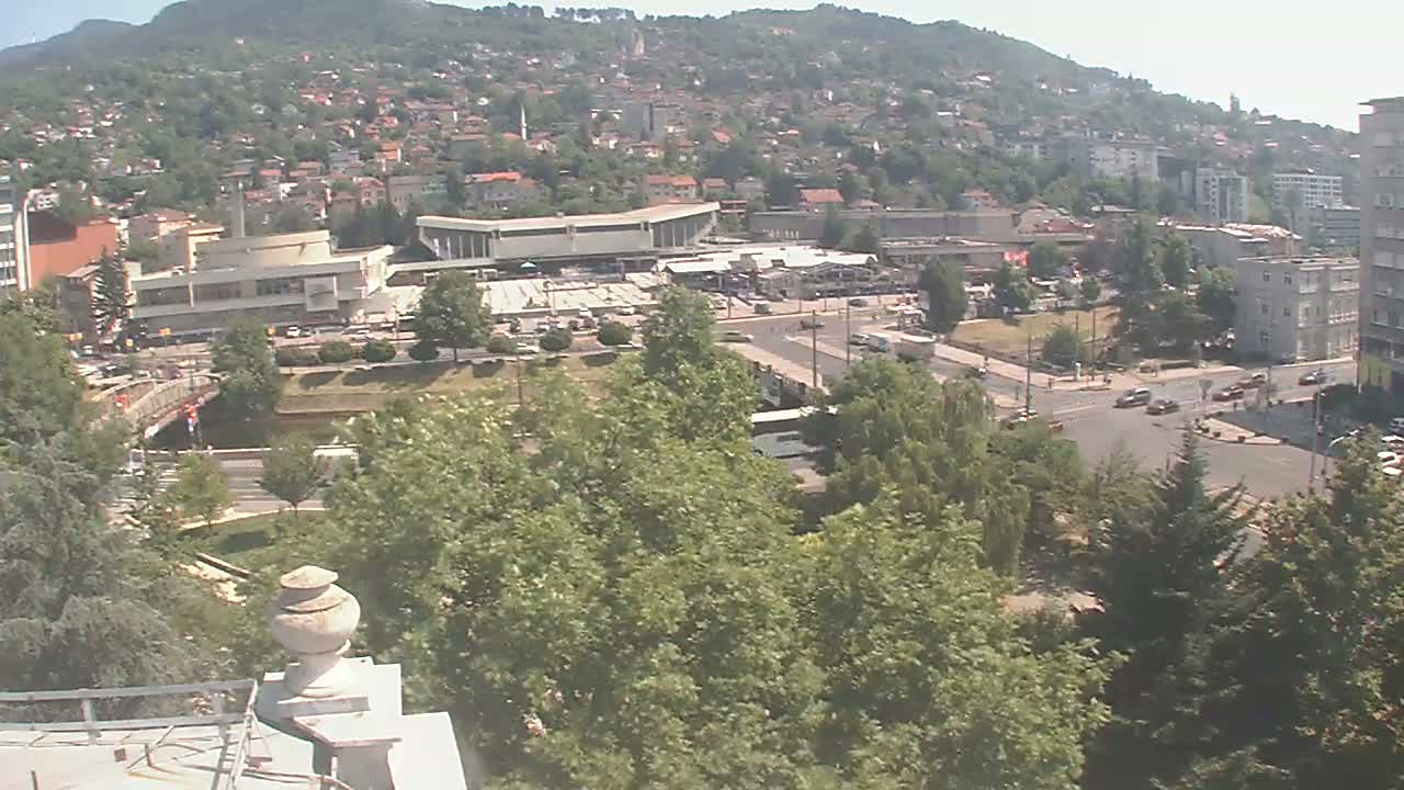 A high-angle view of a city nestled against a hillside on a sunny day, showing buildings, roads, and lush green trees.