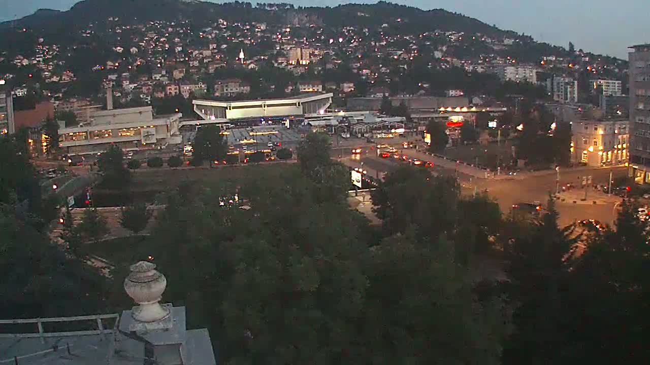 A high-angle view of a city nestled against a hillside on a sunny day, showing buildings, roads, and lush green trees.
