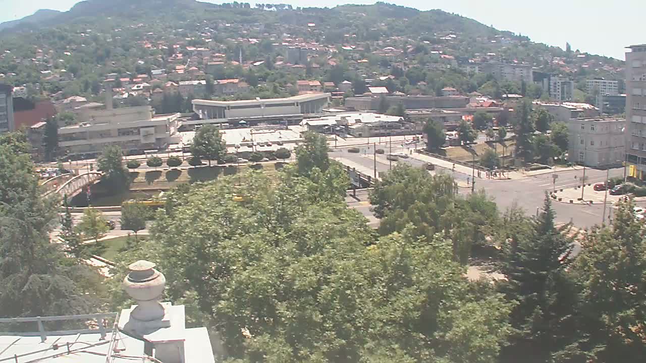 A high-angle, long shot on a sunny day shows a city nestled in a valley, featuring a large plaza, buildings, a bridge over a waterway, and abundant green vegetation.