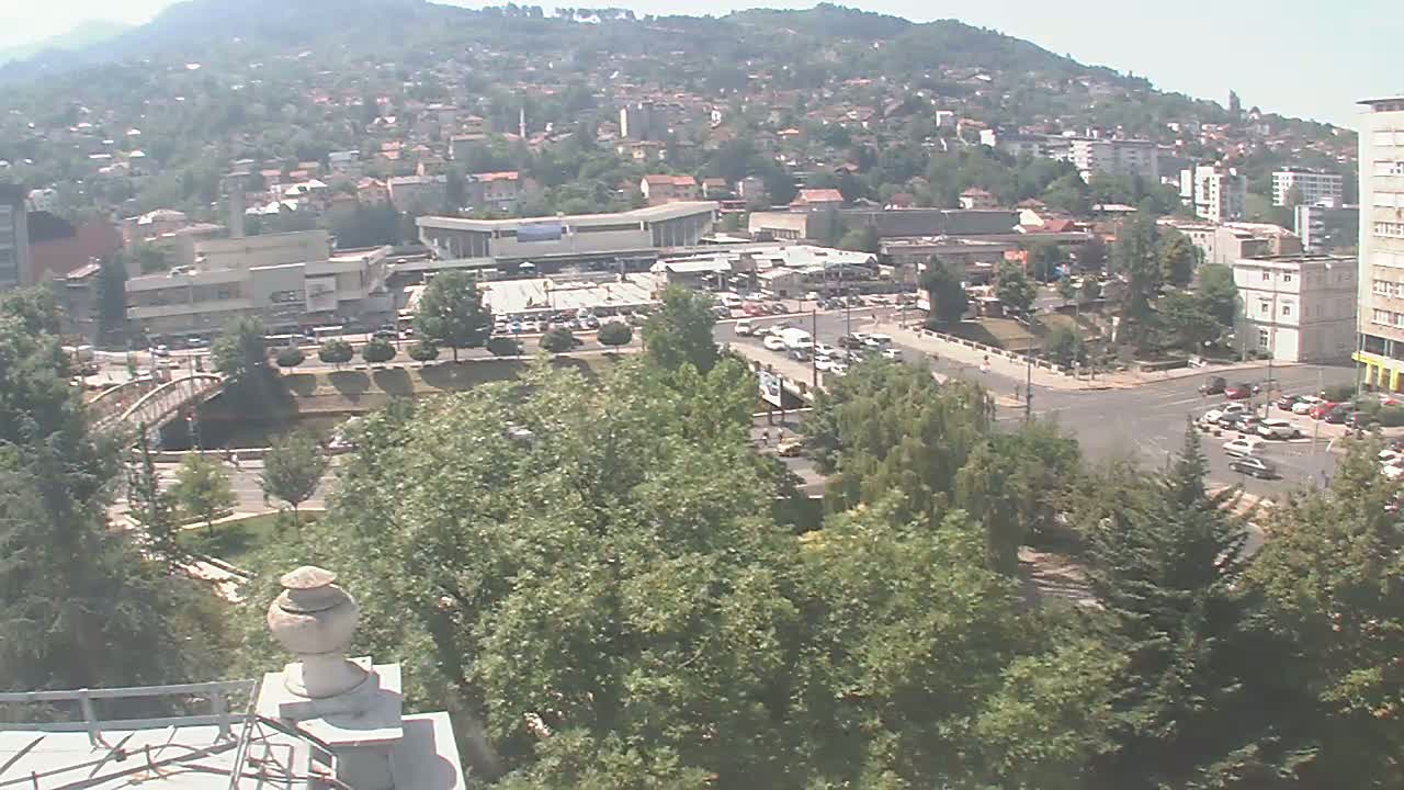 An elevated view of a city on a clear, sunny day, showing a bridge over a waterway, numerous buildings nestled amongst green trees and hills, and several cars traveling on roads.