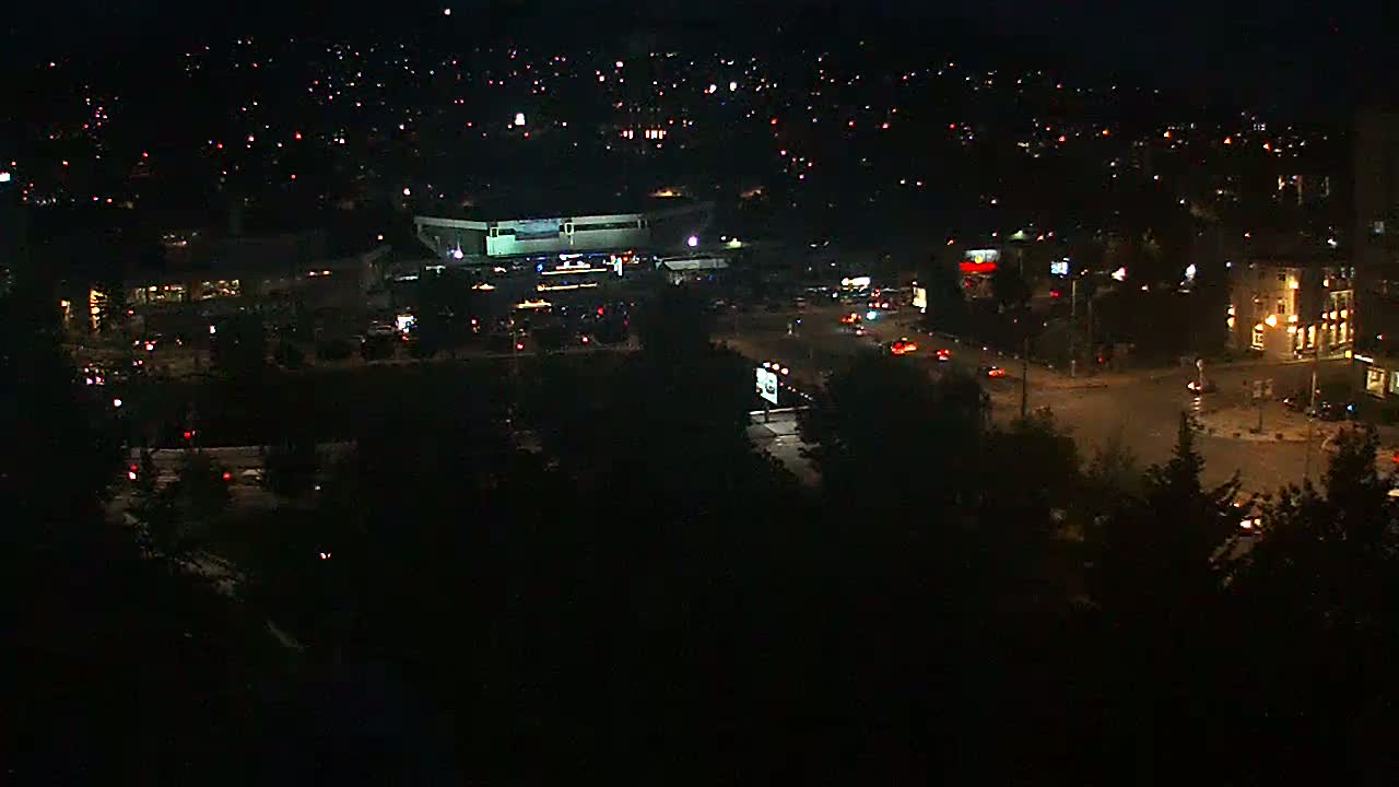A nighttime aerial view shows a city intersection with a large building, surrounded by smaller buildings and trees, under a dark, clear sky.