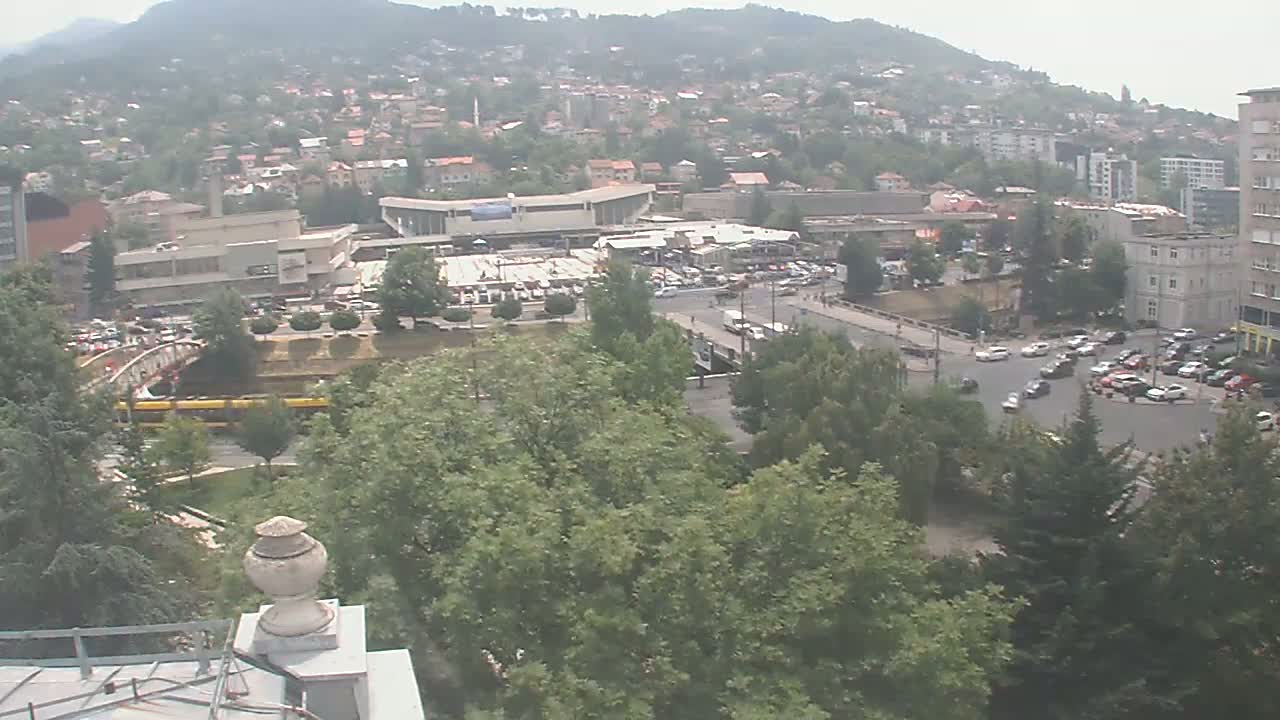 An elevated view of a city on a hazy day shows a large parking lot, buildings, trees, and a tram traveling across a bridge.