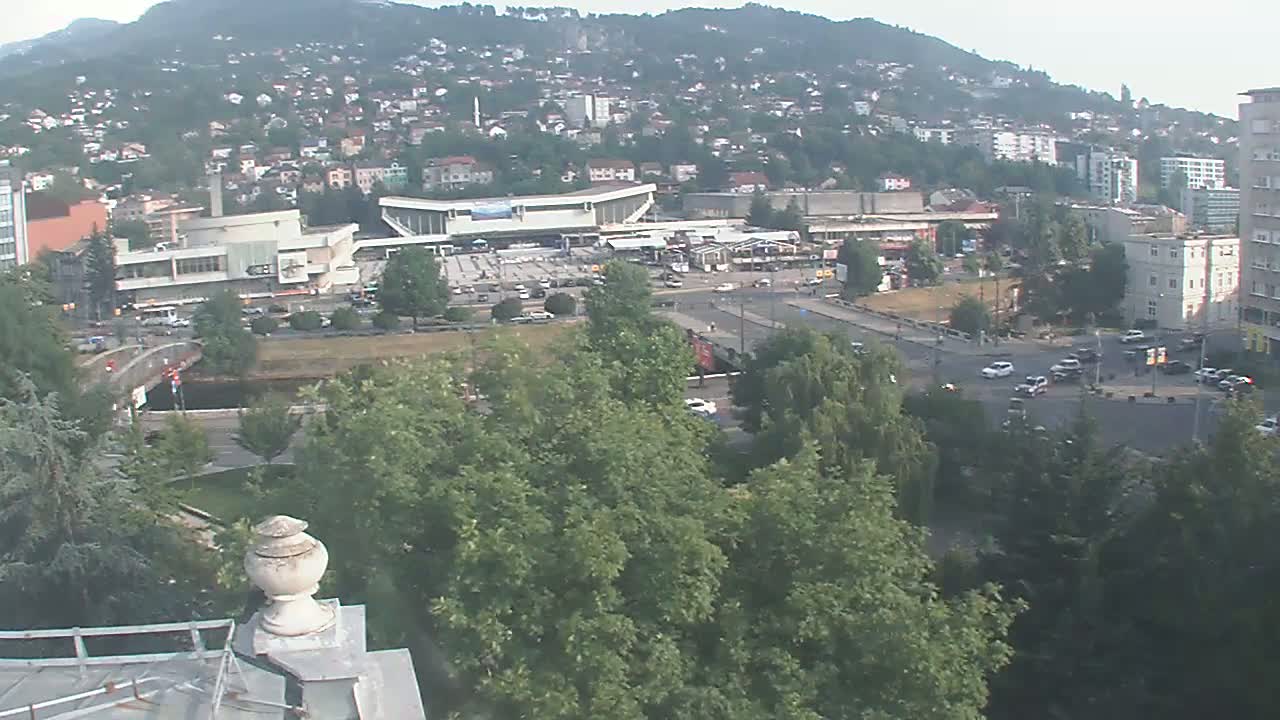 A high-angle view of a city on a hazy day shows a large building complex near a road intersection, surrounded by green trees and hills in the distance.