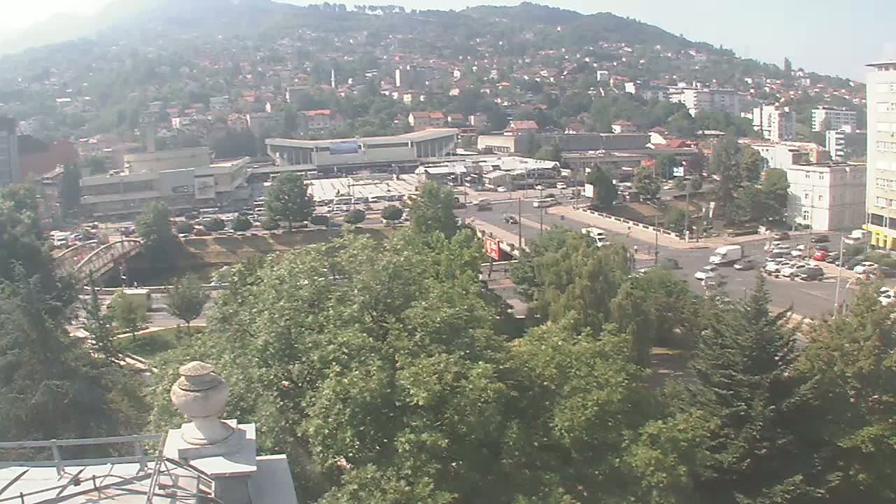 A high-angle view of a city scene on a clear, sunny day, showing a main road intersection, buildings, a bridge, and green spaces, with hills in the background.