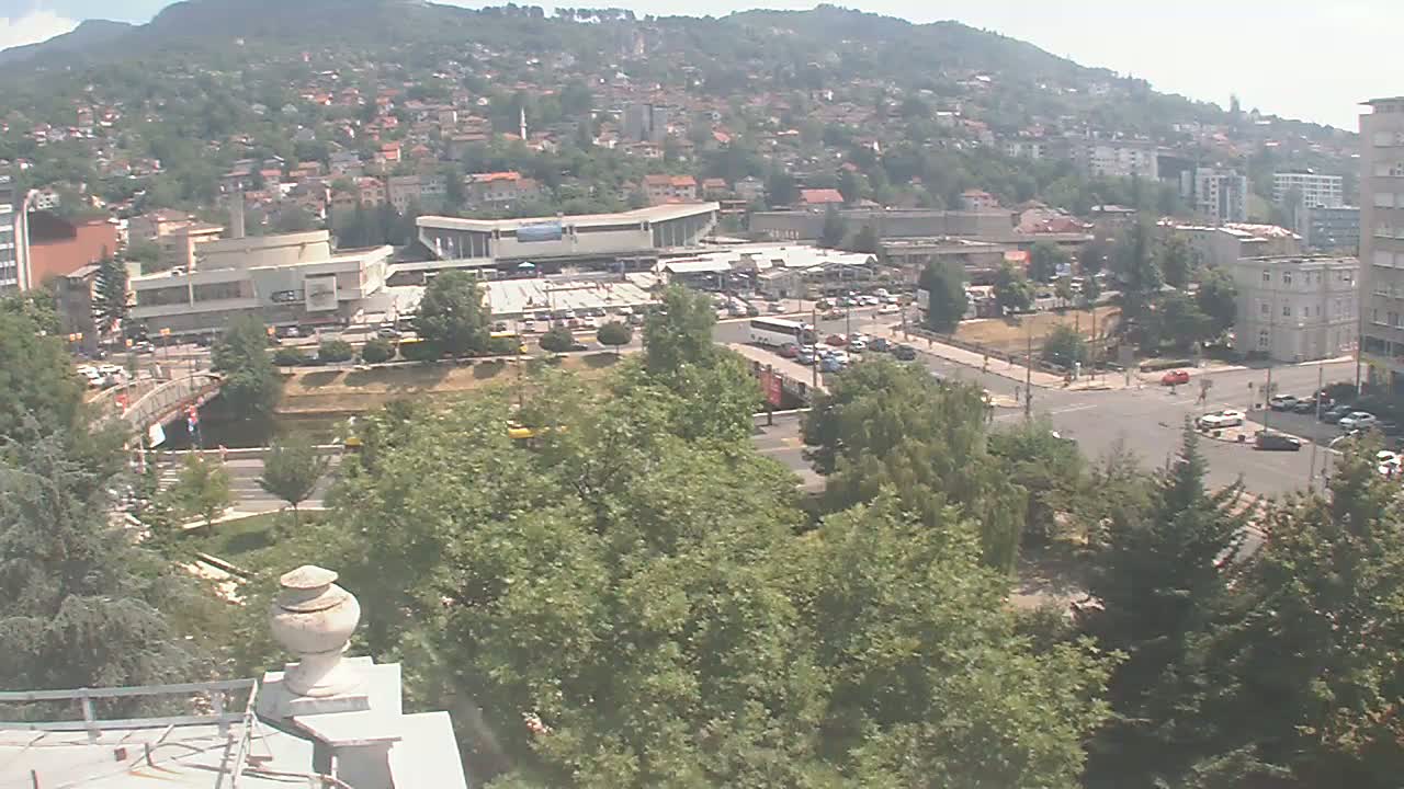 A high-angle view of a city nestled against a hillside on a clear day, showing a main road intersection, a bus station, and numerous buildings and trees.