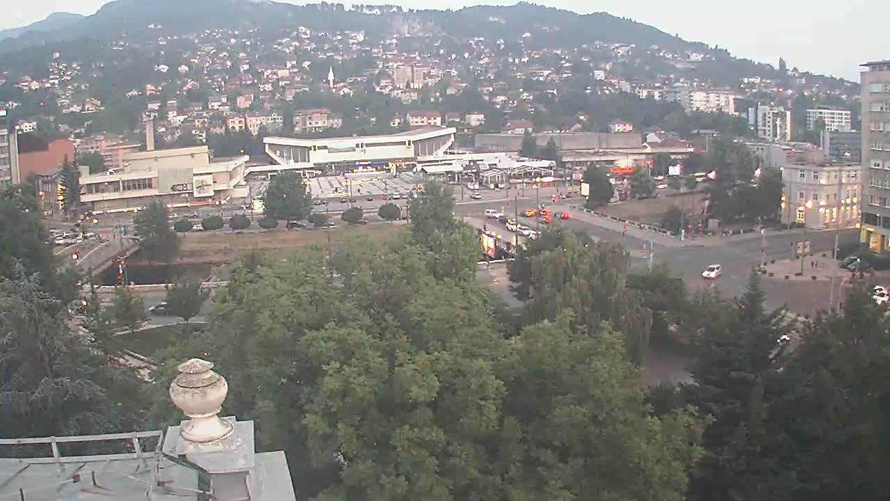 An elevated view of a city at dusk, showing a large building complex near a river, surrounded by lush green trees and hills in the background.