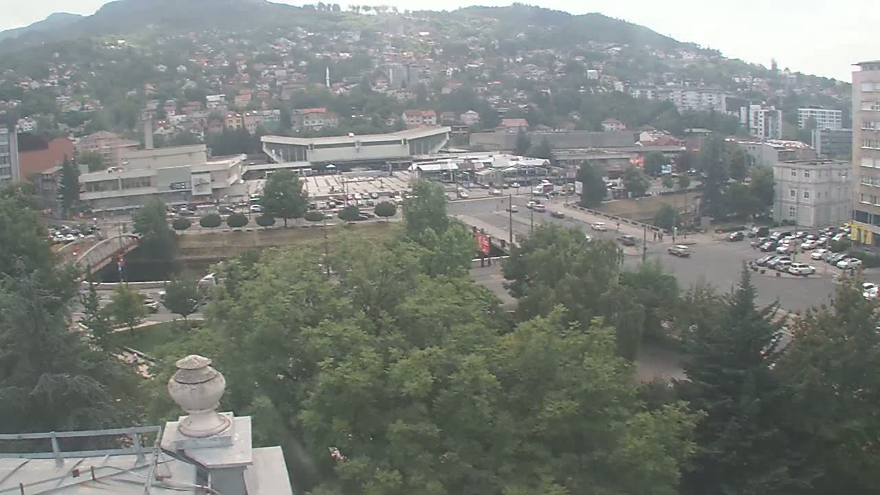 An elevated view of a city on a hazy day shows a large building complex, a bridge over a waterway, and numerous vehicles parked and traveling on streets surrounded by lush green trees.
