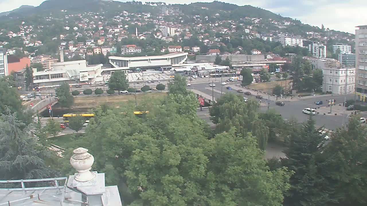 A high-angle view of a city nestled against a hill on a partly cloudy day, showing a bus station, roads, trees, and buildings.