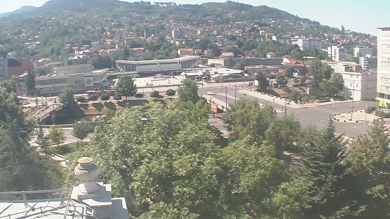 A sprawling town nestled against a hillside on a sunny day, viewed from an elevated position, showing a large plaza, a bridge, and abundant green trees.