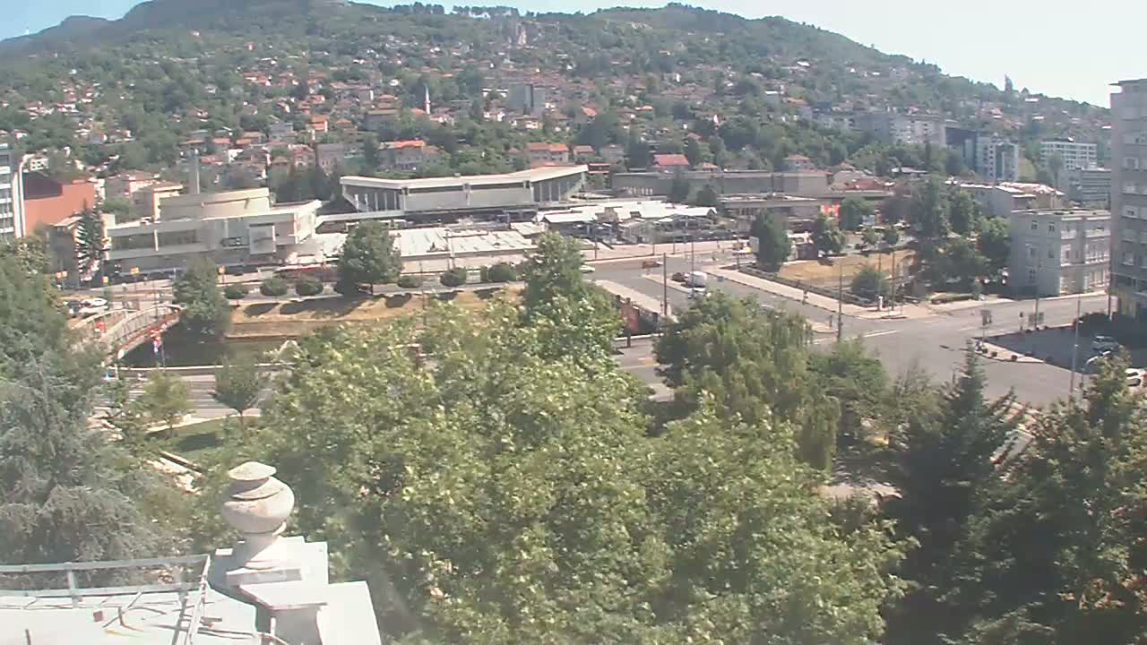 A high-angle view on a sunny day shows a city nestled against a hillside, with a river and bridge visible in the mid-ground and lush green trees in the foreground.