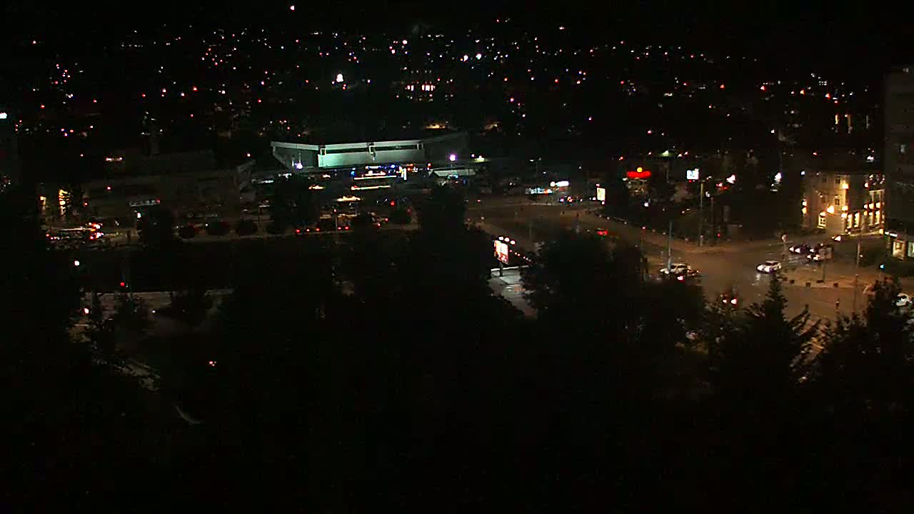 A nighttime aerial view shows a city intersection with sparse traffic, a large building, and surrounding residential areas illuminated by scattered lights.