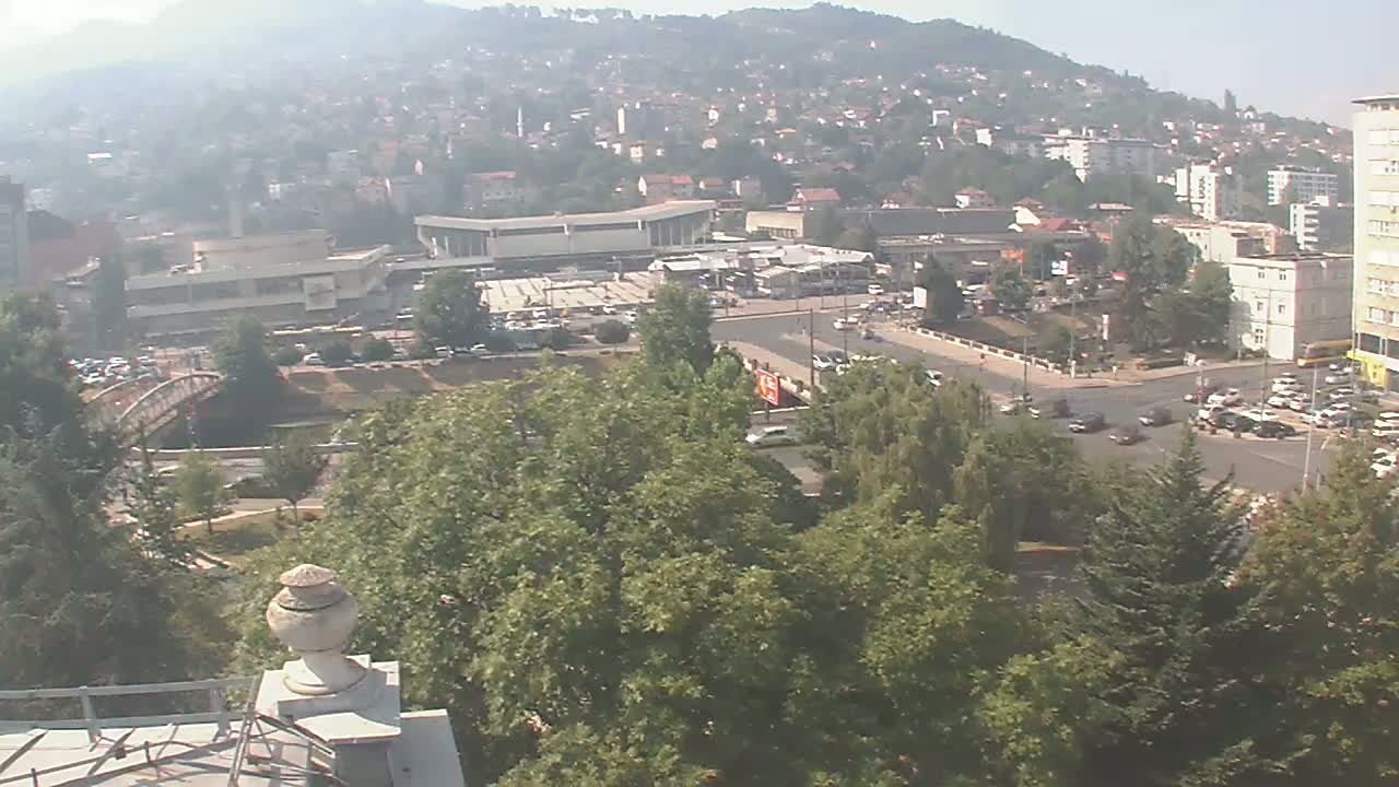 A hazy view of a city reveals a busy intersection with several buildings, a bridge, and a large area of green trees under a clear sky.