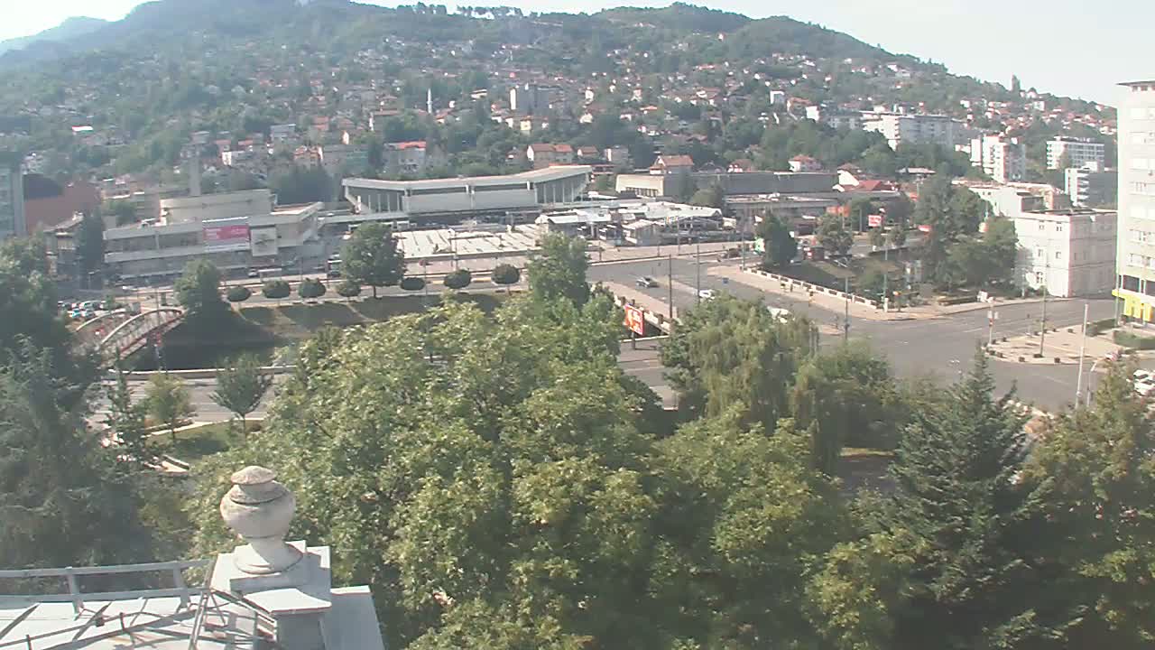 A high-angle view on a sunny day shows a city sprawling across a hillside, with a large plaza and a bridge visible in the foreground, surrounded by lush green trees.