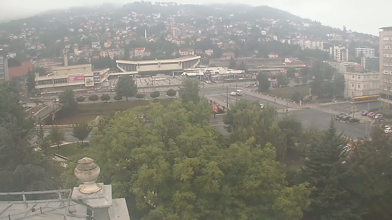 A high-angle view on a sunny day shows a city sprawling across a hillside, with a large plaza and a bridge visible in the foreground, surrounded by lush green trees.