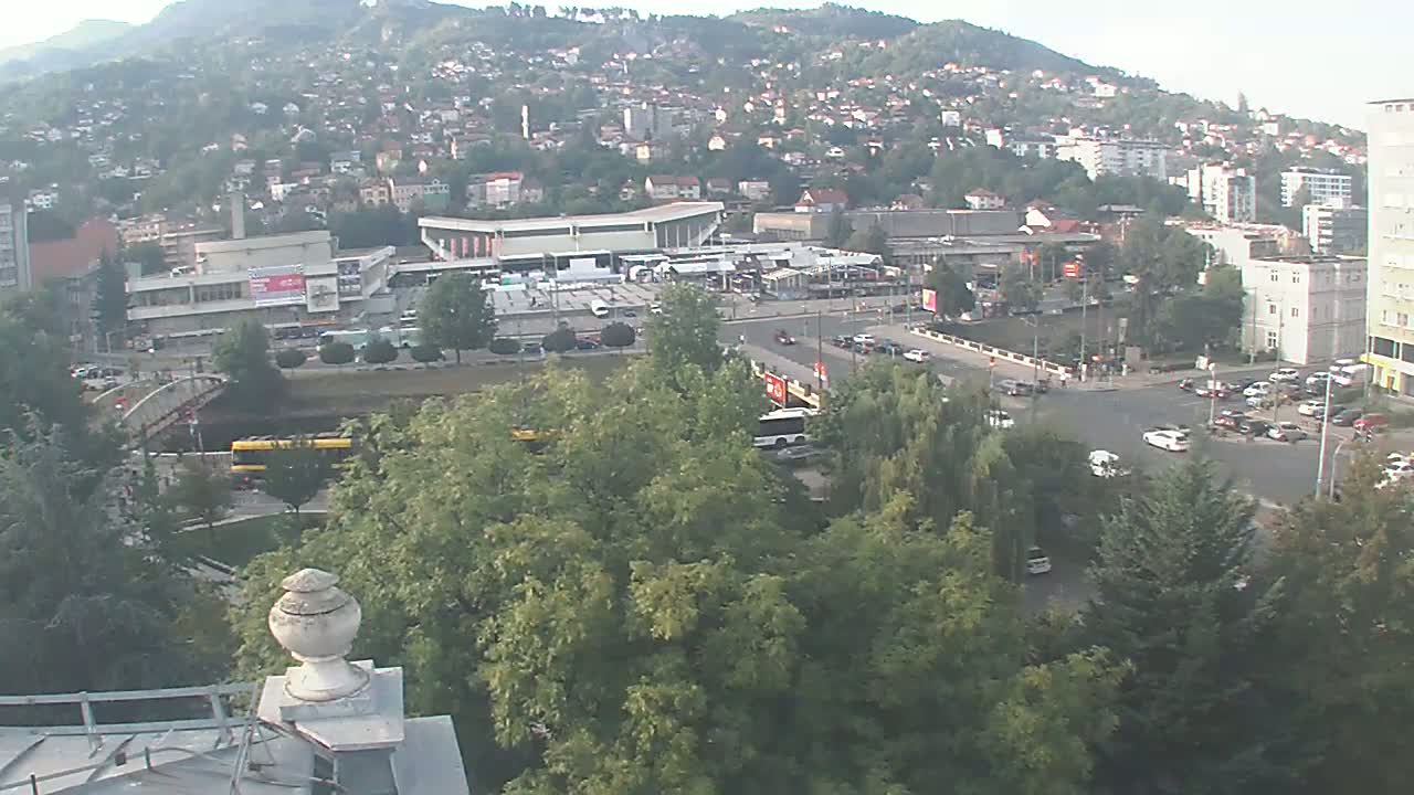 A high-angle view on a sunny day shows a city sprawling across a hillside, with a large plaza and a bridge visible in the foreground, surrounded by lush green trees.
