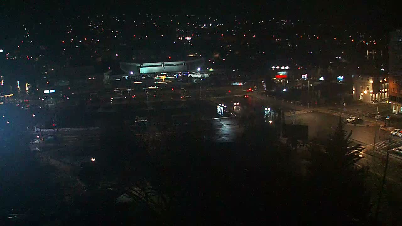 The image captures an elevated nighttime view of a city under wet conditions, with numerous building and vehicle lights illuminating the dark, rain-slicked streets.