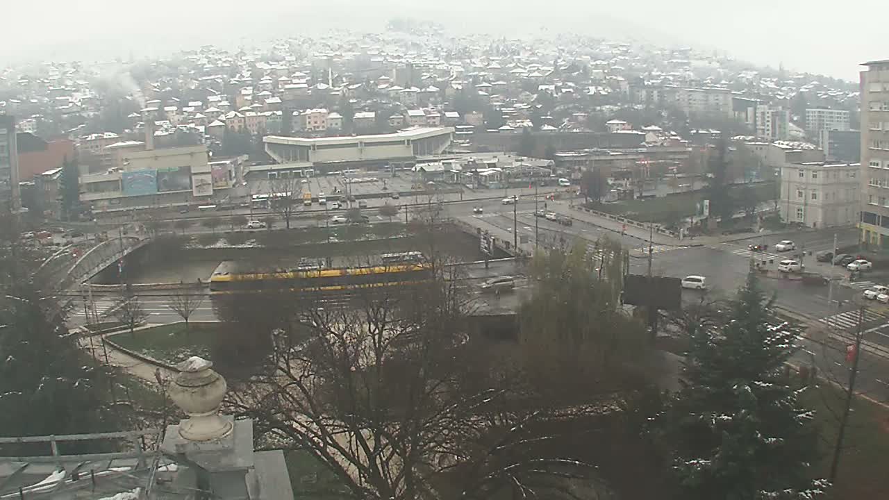 An overcast and misty cityscape reveals light snow on distant hills and evergreens, with a yellow train traveling alongside a river, busy roads, and various urban buildings.