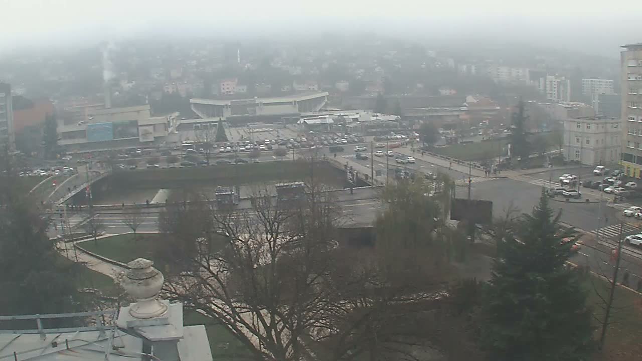 An elevated, hazy view of a city on an overcast day shows extensive road networks with vehicles, a multi-level bridge system, dense buildings including a distant stadium, and scattered dormant trees.