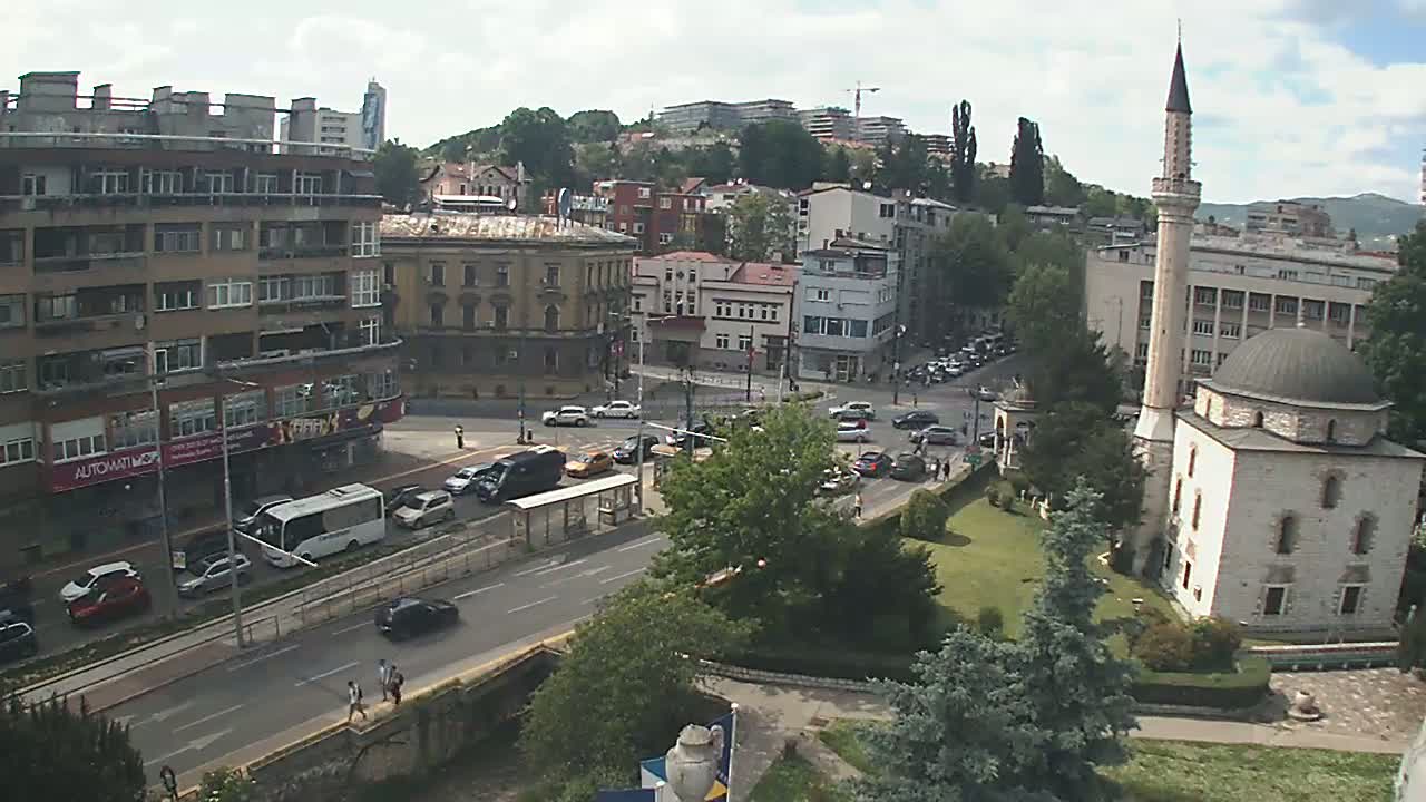 A high-angle view of a city street scene on a partly cloudy day, featuring a mosque, buildings, traffic, and green spaces.