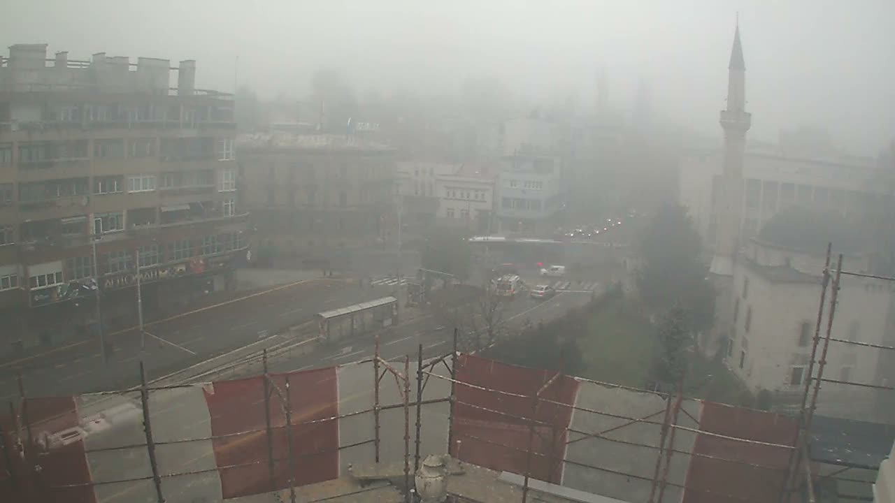 An urban street scene, viewed from behind scaffolding with red and white netting, reveals cars and buildings, including a mosque minaret, all heavily obscured by dense fog or smog.