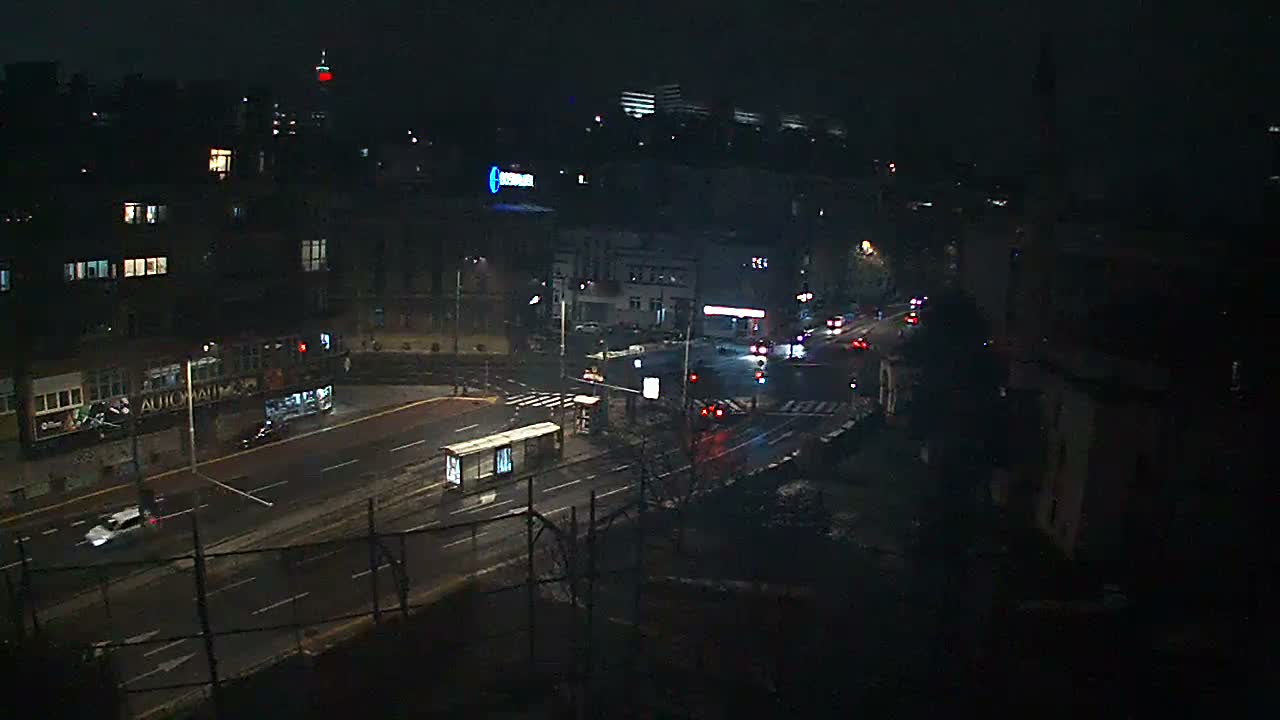 A dark city street at night shows vehicles driving on wet pavement, illuminated by streetlights, building lights, and car headlights and taillights.