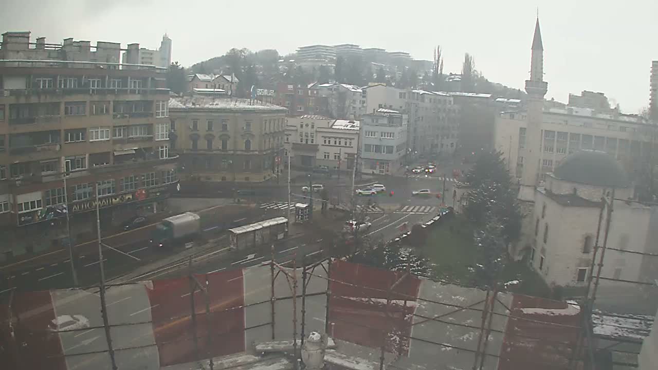 An overcast winter cityscape, lightly dusted with snow on rooftops and trees, depicts a busy road with traffic, diverse buildings, and a prominent mosque, all under a hazy sky.