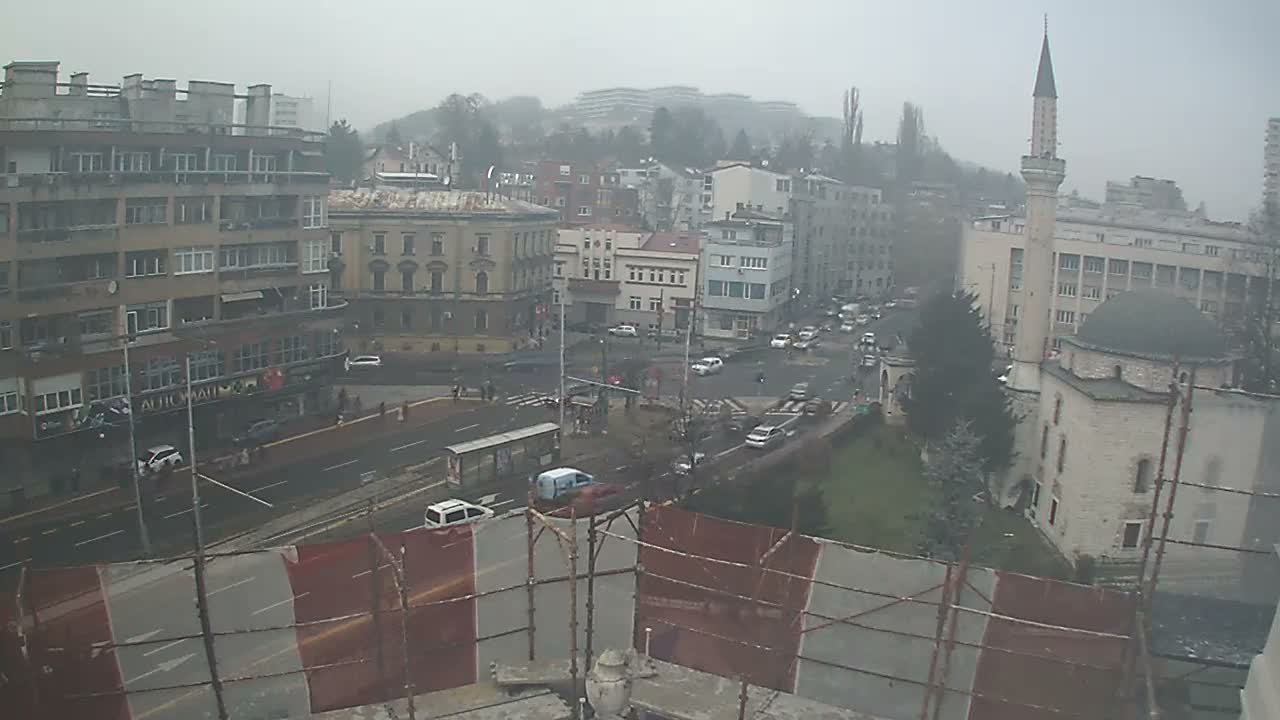 A wide, hazy and overcast view of an urban area shows a main road with active vehicle traffic, a prominent mosque with a minaret, and various other buildings set against partially obscured distant hills.