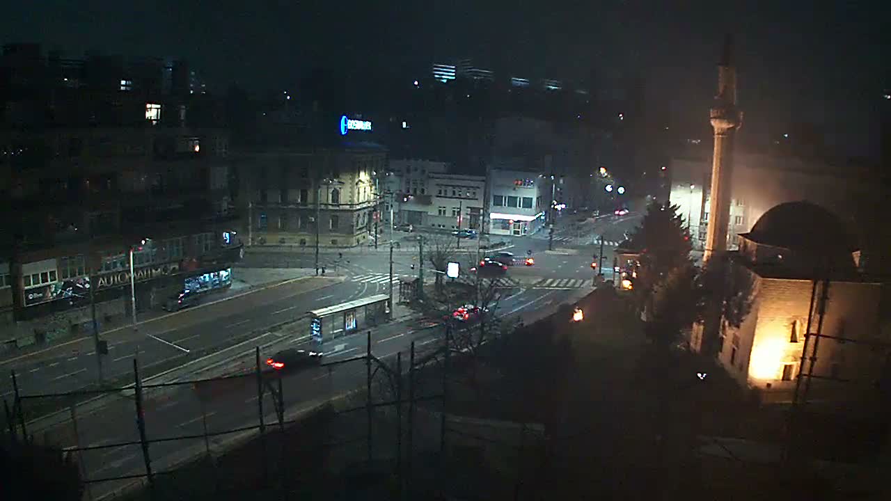 An elevated nighttime view shows a city intersection with moving cars and illuminated buildings, including a prominent mosque with its minaret, under slightly hazy conditions.