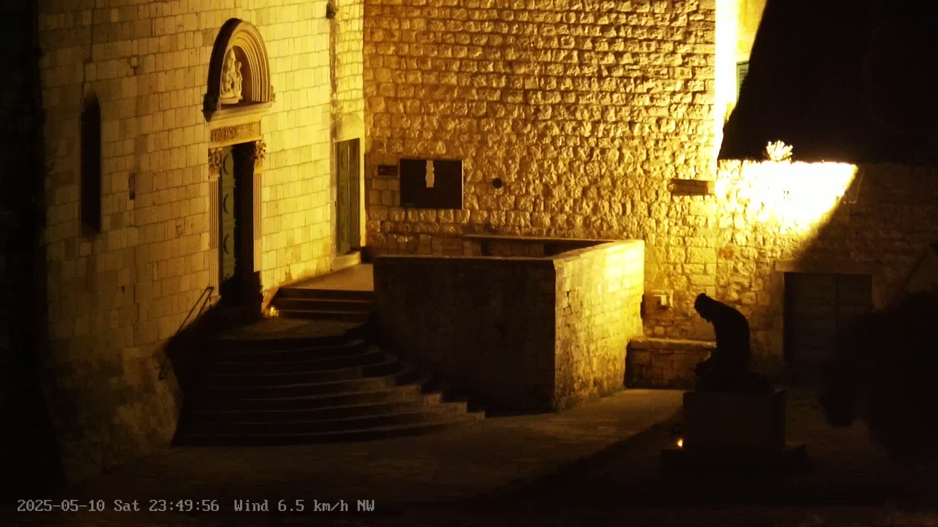 A stone building is illuminated at night by artificial light, showcasing a doorway with steps and a shadowed figure sitting on a low structure.