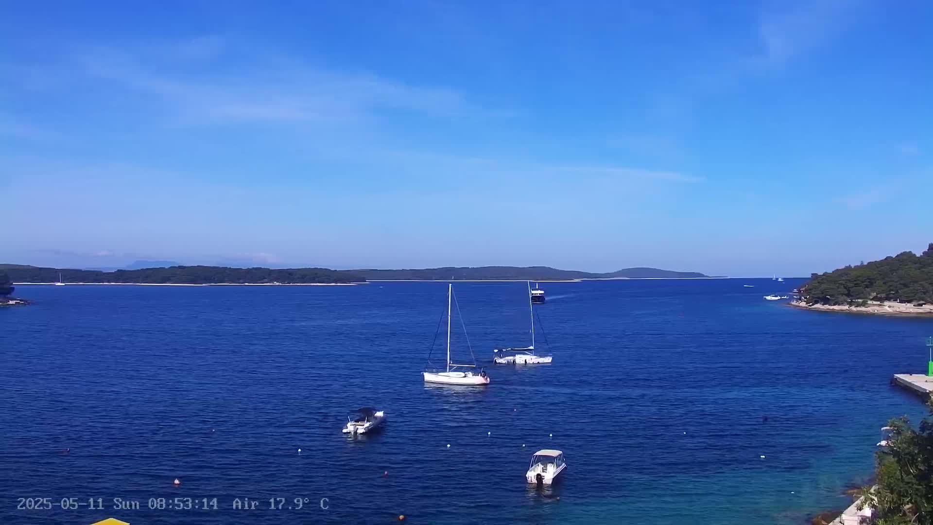 Several sailboats and motorboats are anchored in a calm, blue sea on a sunny day, with a distant shoreline visible under a clear sky.