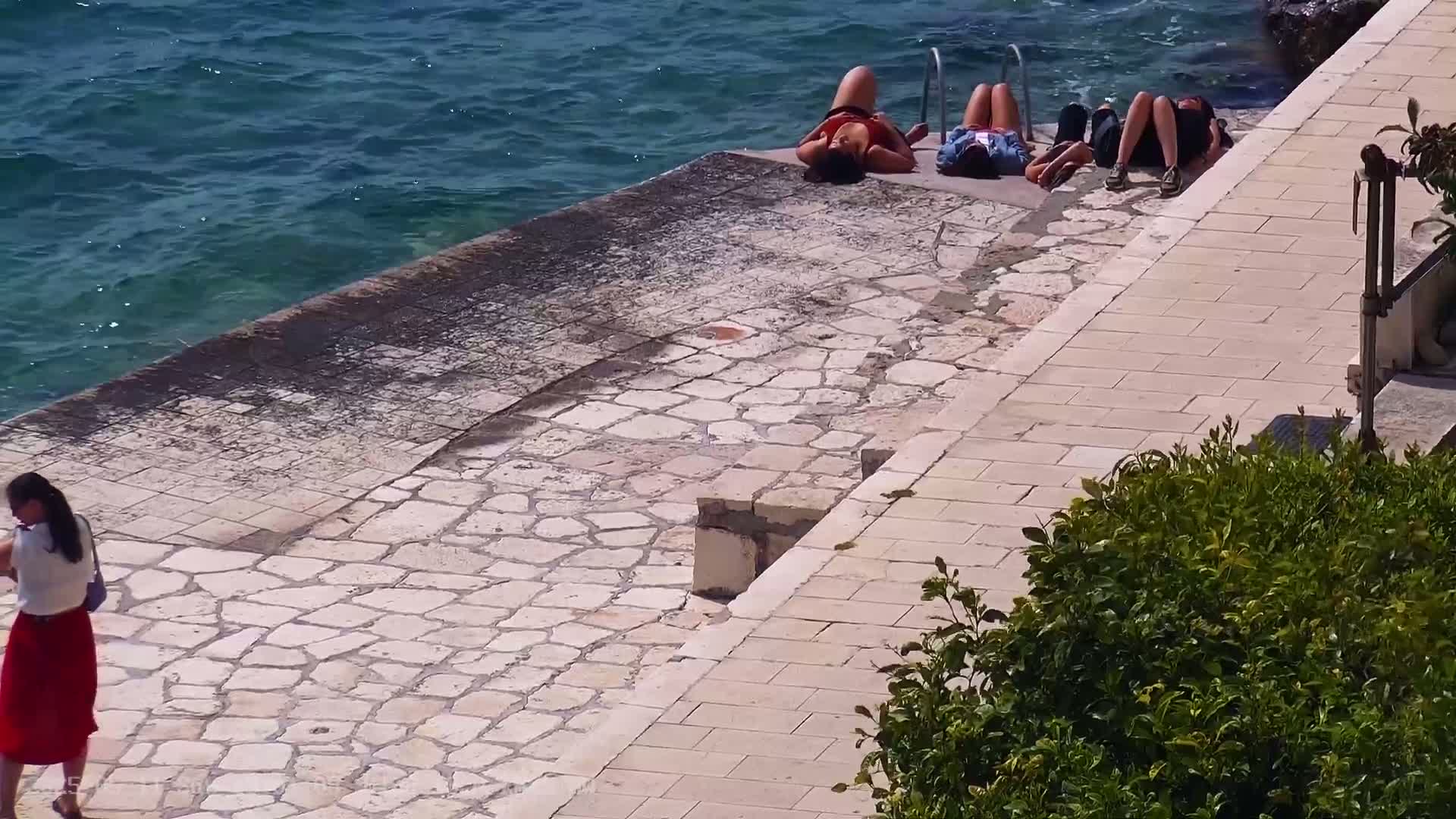 Three people sunbathe on a stone surface next to a calm body of water, while a person in a red skirt walks along a paved path.
