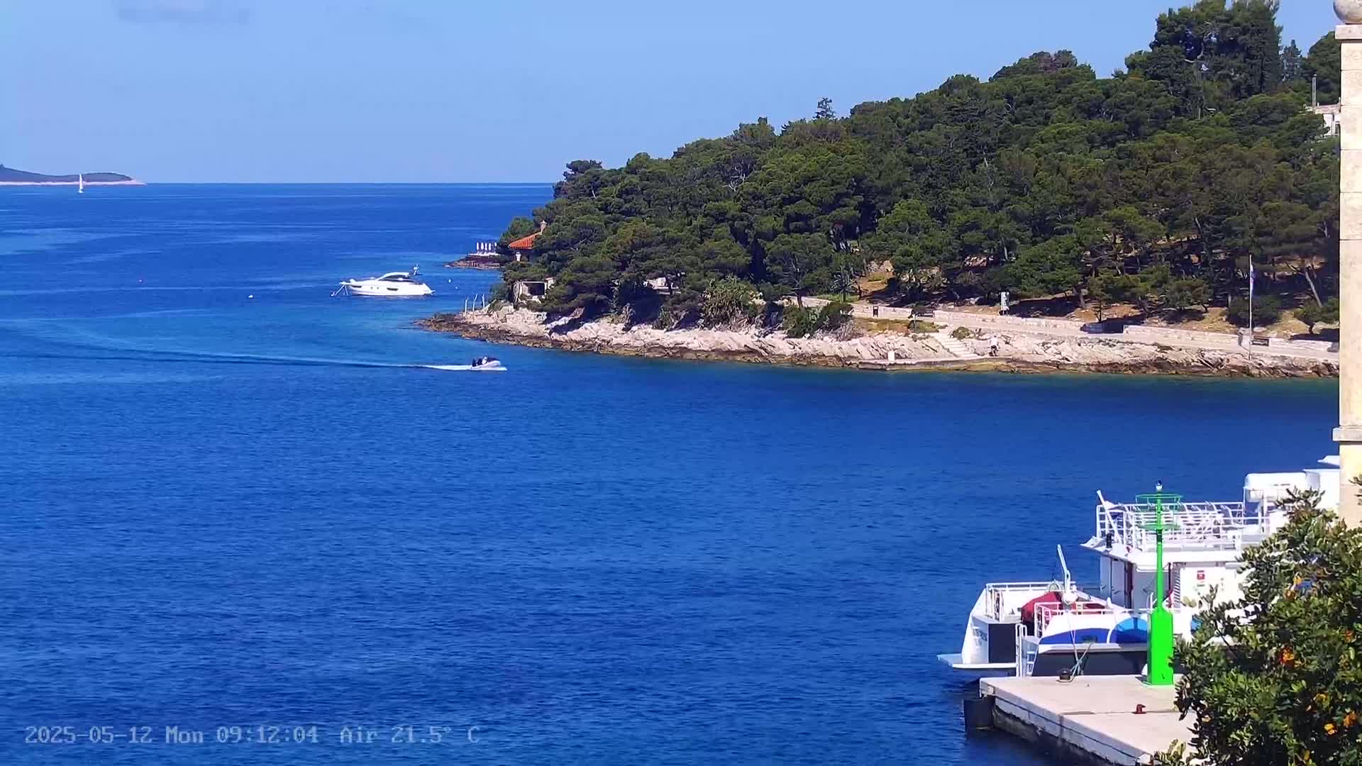 A sunny, clear day shows a calm blue sea with several boats, a small landmass with green trees, and a larger body of land in the distance.