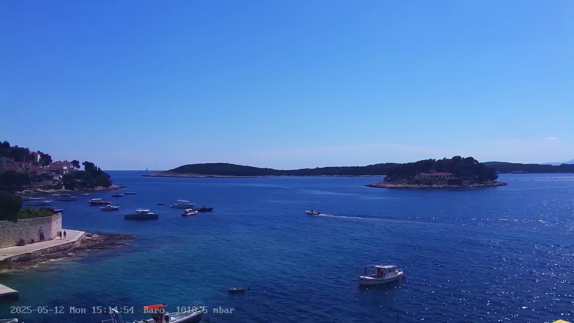 A calm, sunny day shows a clear blue sea dotted with small boats near a shoreline with buildings, and a small island with a house in the distance.
