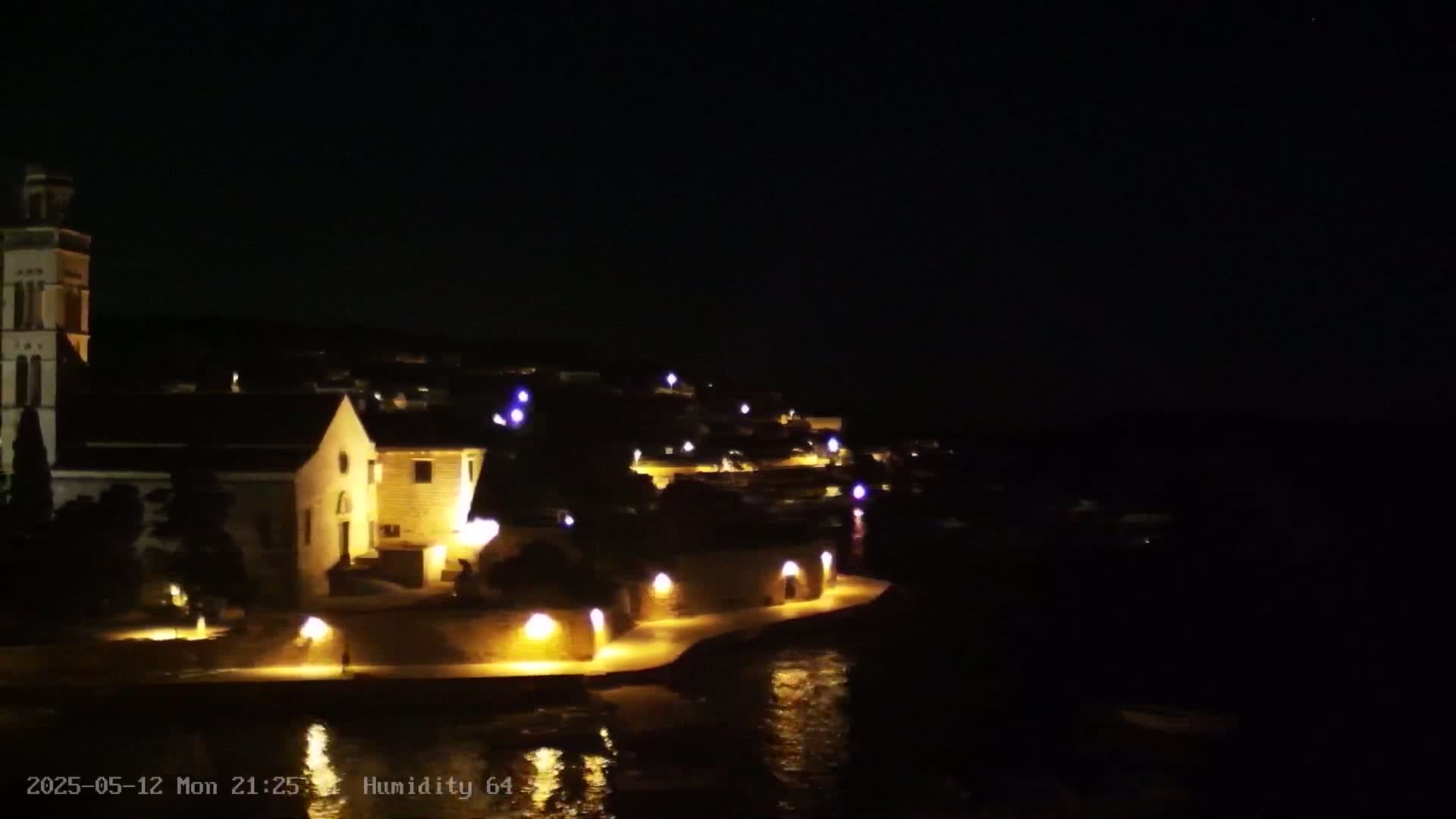 A nighttime view of a coastal town with a church, illuminated buildings, and calm water reflecting the lights.