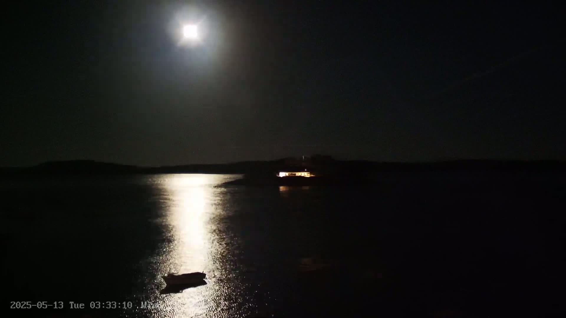 A bright moon illuminates a calm body of water at night, reflecting its light onto the surface where a small boat is visible, and a building with lights is seen on a distant shore.