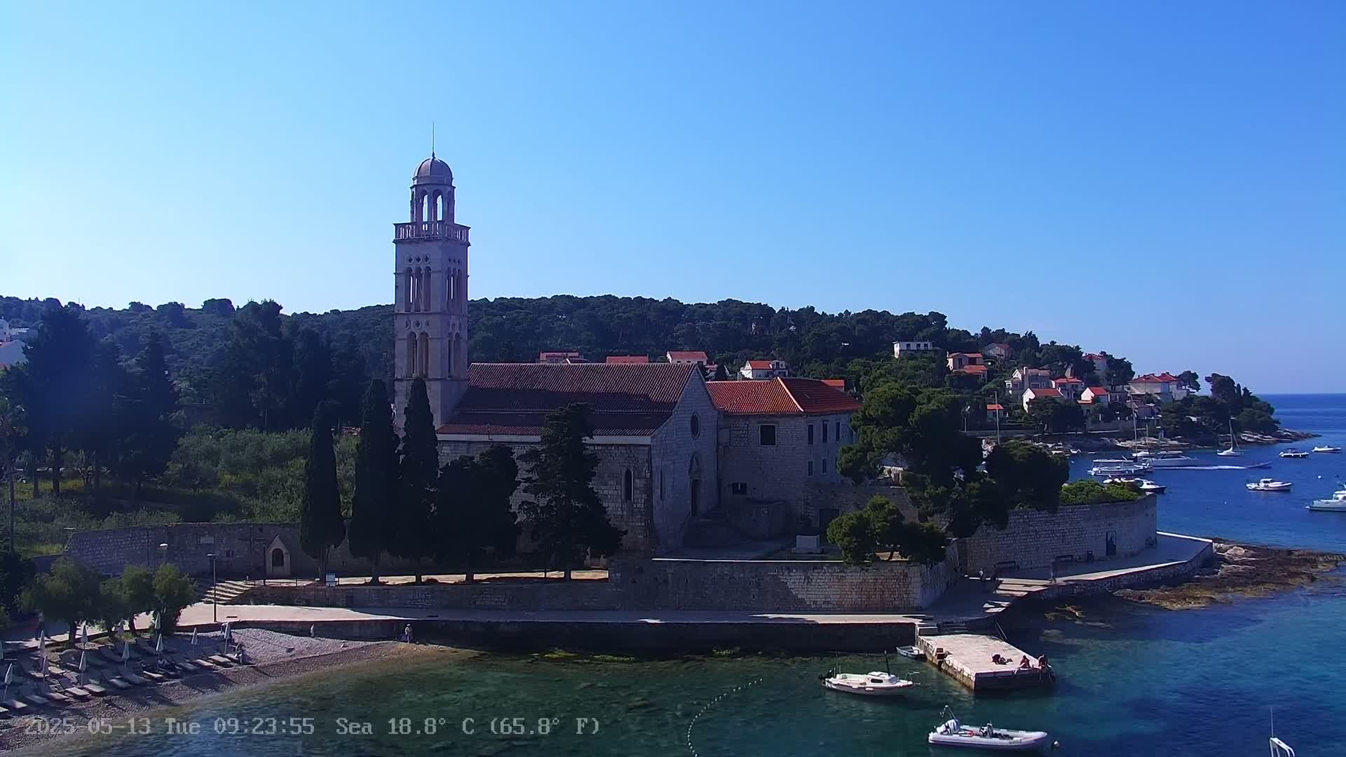 A stone church with a tall bell tower sits on a coastline with clear blue water, boats, and a small beach under a bright, sunny sky.