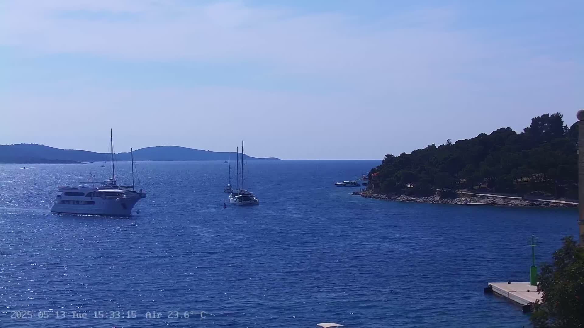 Several boats are sailing on a calm, blue sea near a rocky shoreline with green trees under a clear, bright sky.